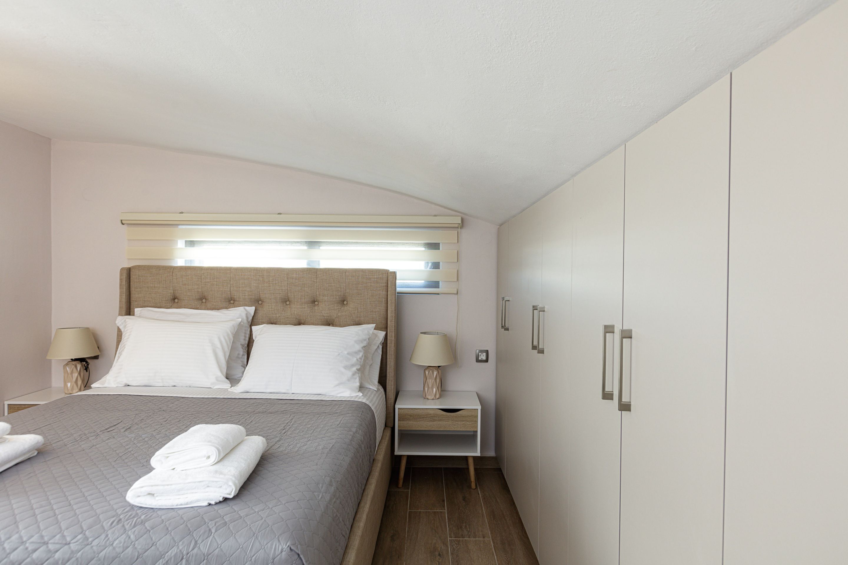 A neatly made bed with a quilted cover, two bedside lamps, a nightstand, and a wardrobe with two doors.