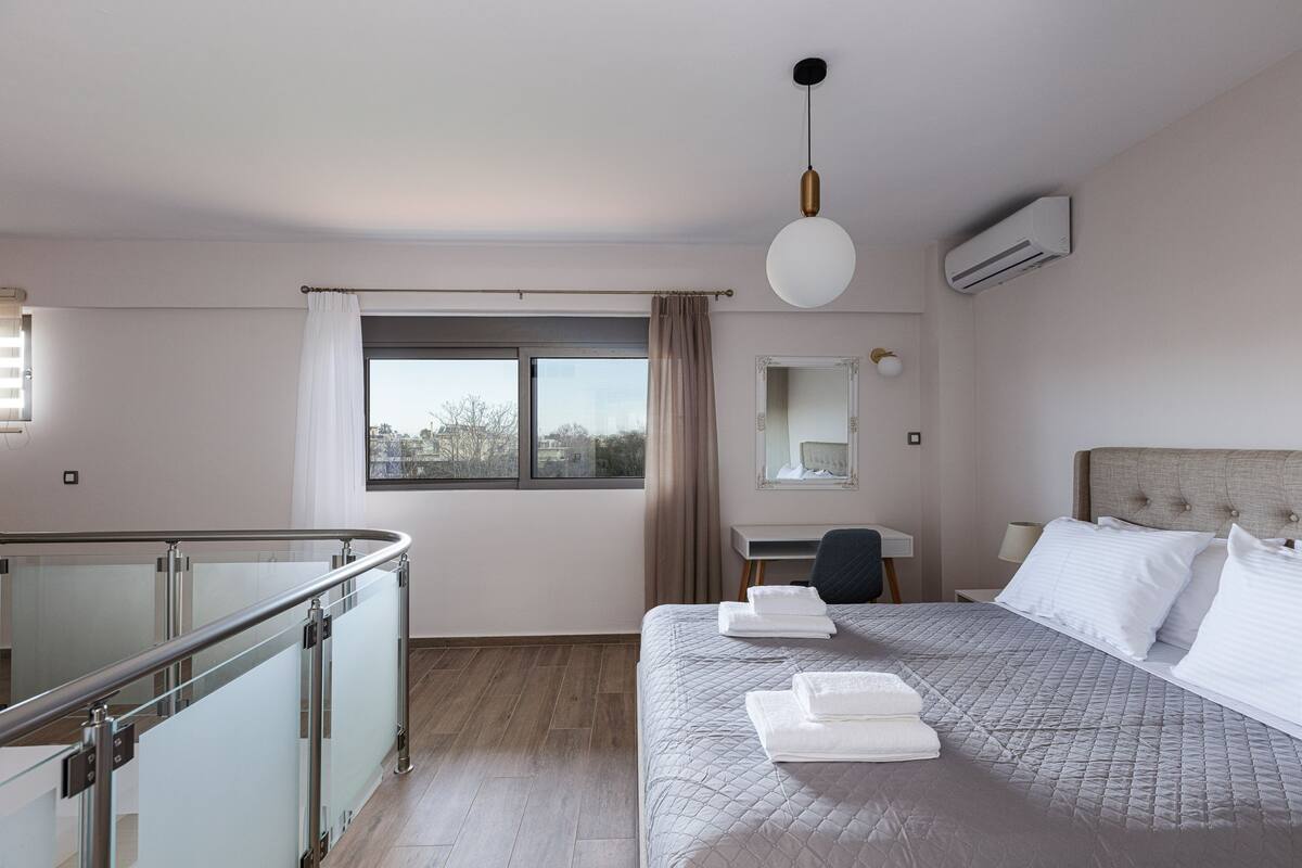 A hotel room with a bed, a desk, a chair, a window with curtains, and an air conditioning unit.