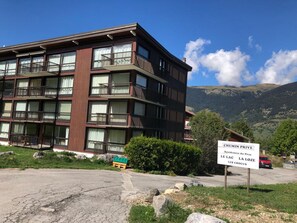 A multi-story building with balconies, a sign indicating 'CHEMIN PRIVE Residences du Praz', and a mountainous backdrop.