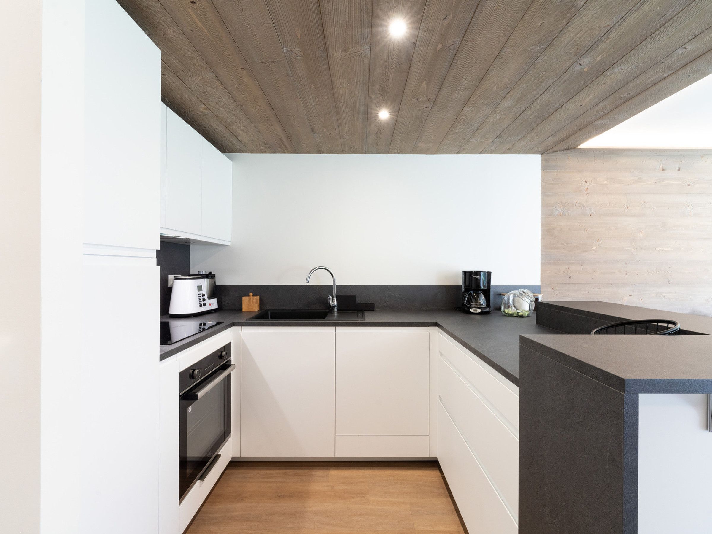 A modern kitchen with white cabinets, a black countertop, a wooden ceiling, and a built-in oven.