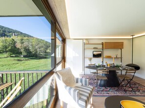 Un salon moderne doté d’une grande fenêtre donnant sur un paysage verdoyant, un coin repas avec une table dressée pour deux personnes, et une cuisine équipée d’un téléviseur et de meubles en bois.