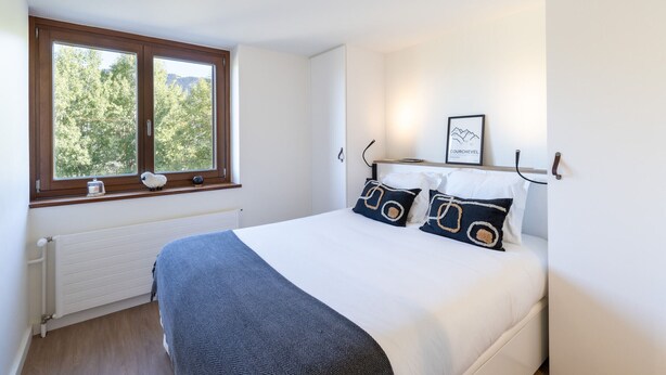 A bedroom with a large window, a bed with white bedding and patterned pillows, a radiator, and a framed picture on the wall. - Renovated apartment near slopes with Wi-Fi for families (Courchevel)