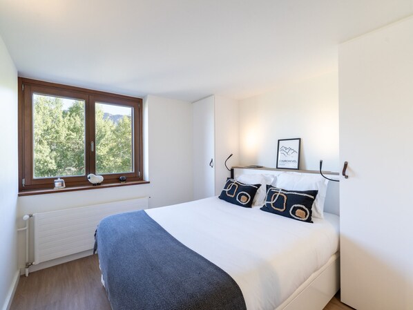 A bedroom with a large window, a bed with white bedding and patterned pillows, a radiator, and a framed picture on the wall. - Renovated apartment near slopes with Wi-Fi for families (Courchevel)