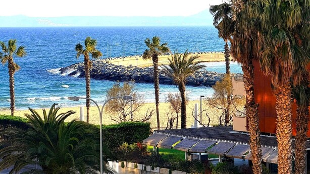 A coastal view with a rocky beach, palm trees, and a building with solar panels. - Comfortable apartment, 50m from beach, 4 people, terrace, TV, washing machine (Roses)