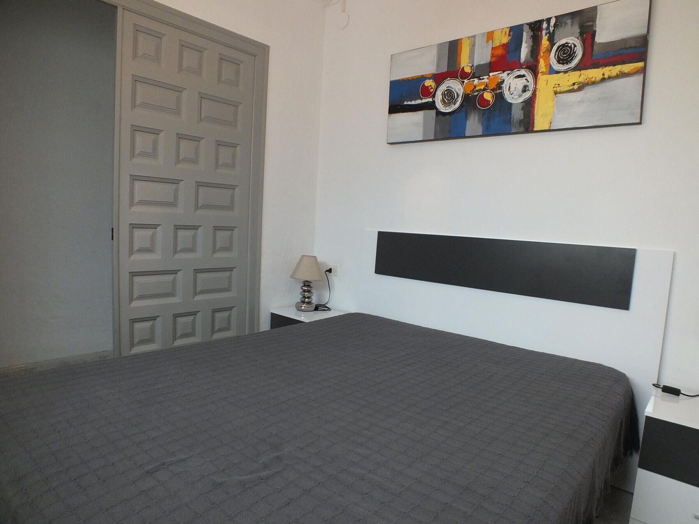 A bedroom with a bed, a nightstand, a lamp, a door, and a painting on the wall.