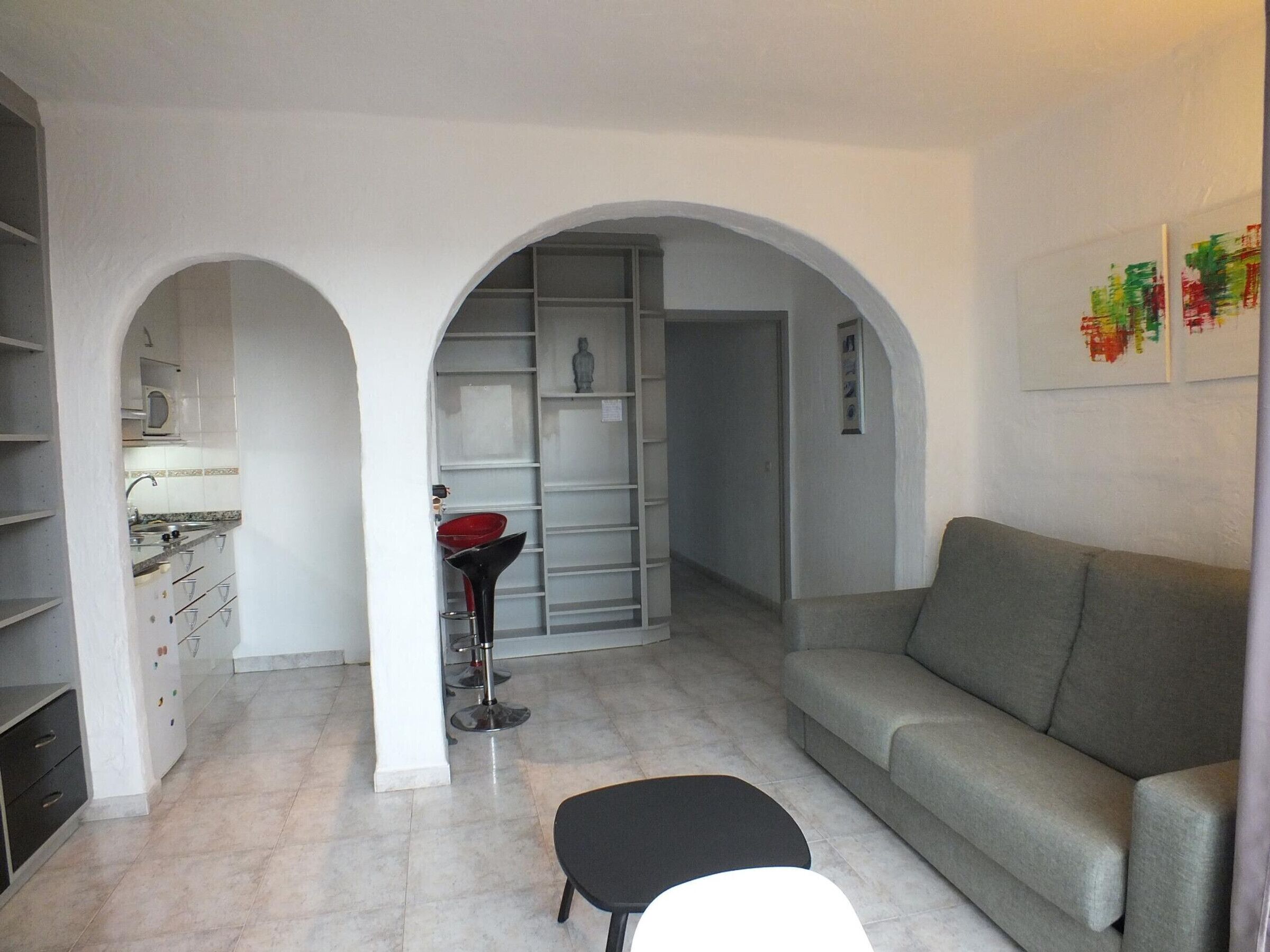 A living room with a grey sofa, a black ottoman, and a kitchen area with white cabinets and a red chair.