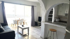 A modern living room with a sofa, a dining table, and chairs, a kitchen area with a counter and cabinets, and a large window with curtains. - Comfortable apartment, 50m from beach, 4 people, terrace, TV, washing machine (Roses)