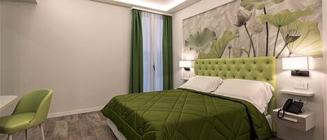 Habitación de hotel con una cama y una silla verdes, un cuadro de hojas y una mesita de noche con un teléfono.