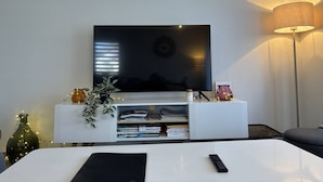 A modern living room with a flat-screen TV mounted on a white entertainment unit, a floor lamp, and a small plant. - Peaceful Mountain Escape: 2BR Apartment, Terrace, Close to Ski, Free Parking, WiFi (Villard-de-Lans)