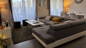 A modern living room with a grey sofa, a round mirror, a clock on the wall, and a dining area with chairs and a table. - Peaceful Mountain Escape: 2BR Apartment, Terrace, Close to Ski, Free Parking, WiFi (Villard-de-Lans)