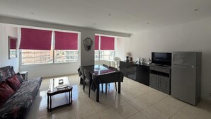 A modern living room with a sofa, dining table, and kitchen area including a refrigerator and TV. - Charming 1-bedroom apartment with pool & garden in Roses Santa-Margarita (Roses)