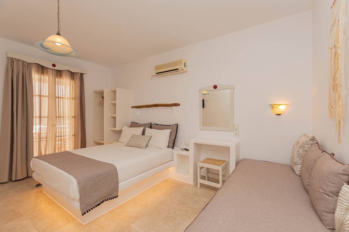 Un dormitorio con una cama, mesitas de noche, un taburete pequeño, un espejo y un equipo de aire acondicionado.