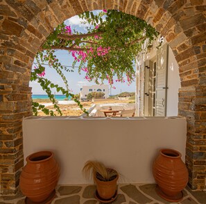 A stone archway with a view of a beach and a white building with a door. There are two terracotta pots on the ground. - Studio Strouvalis (Naxos)