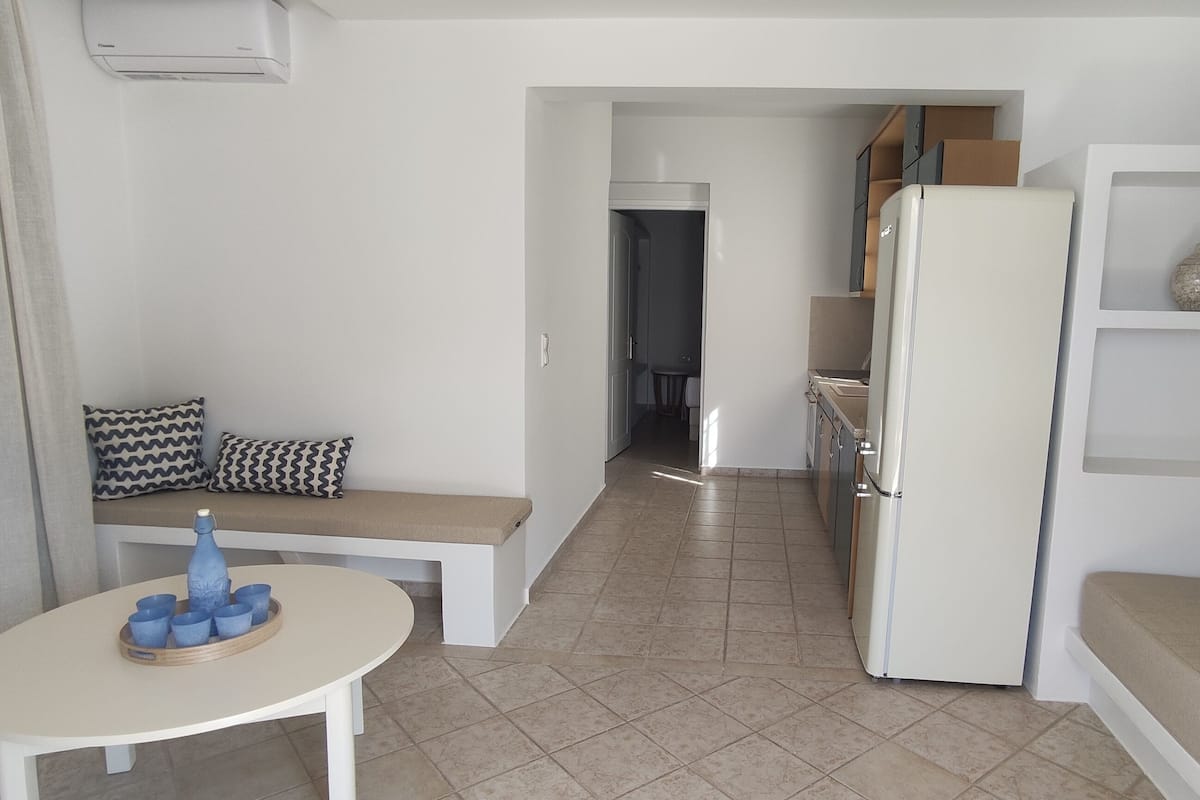 Un espacio habitable compacto con kitchenette, una zona de comedor con mesa y sillas, y un rincón de estar con sofá y cojines.