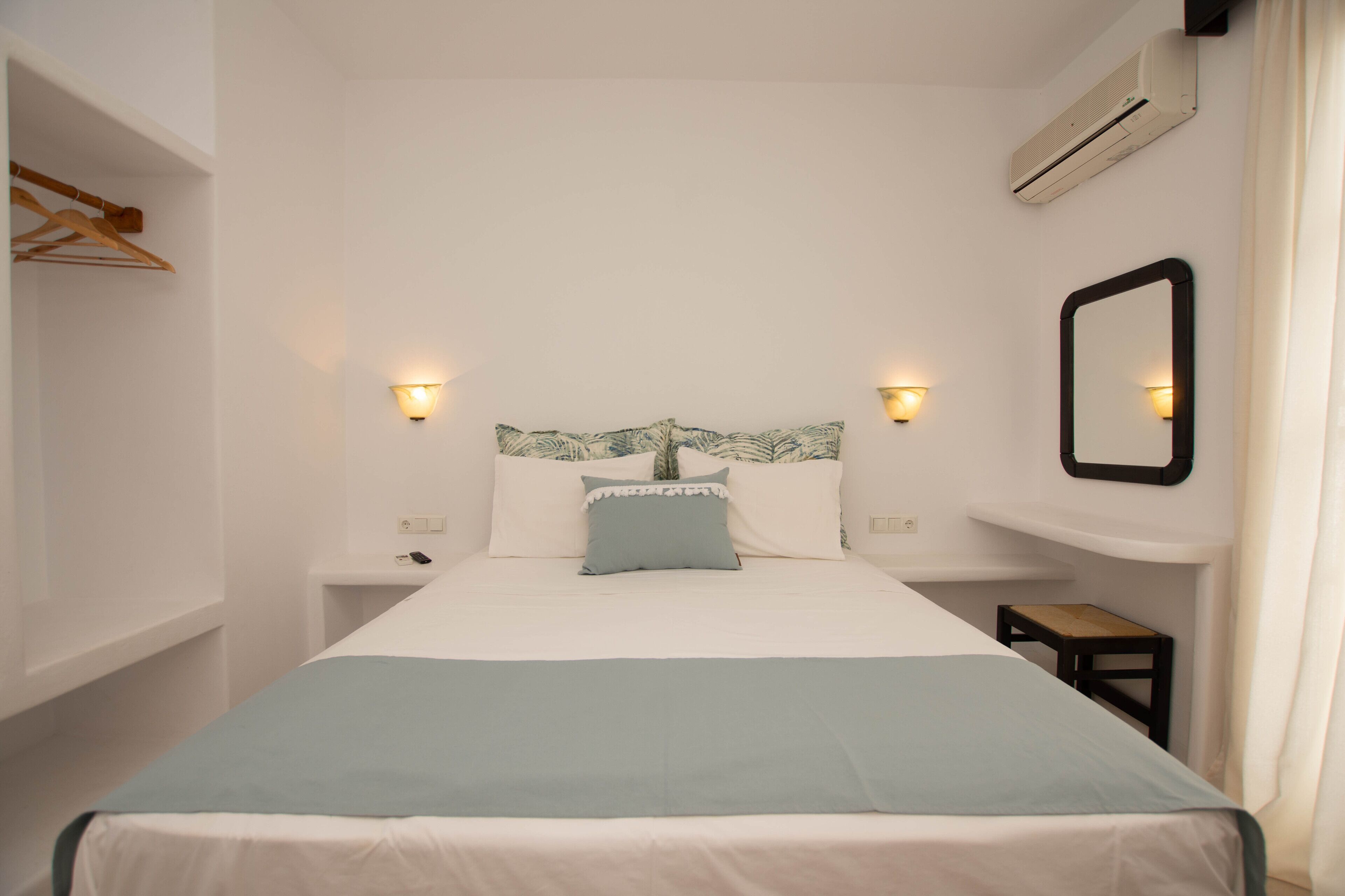 Un lit bien fait, recouvert d’une couvre-lit bleue, avec deux lampes de chevet, un miroir fixé au mur et un petit tabouret en bois.