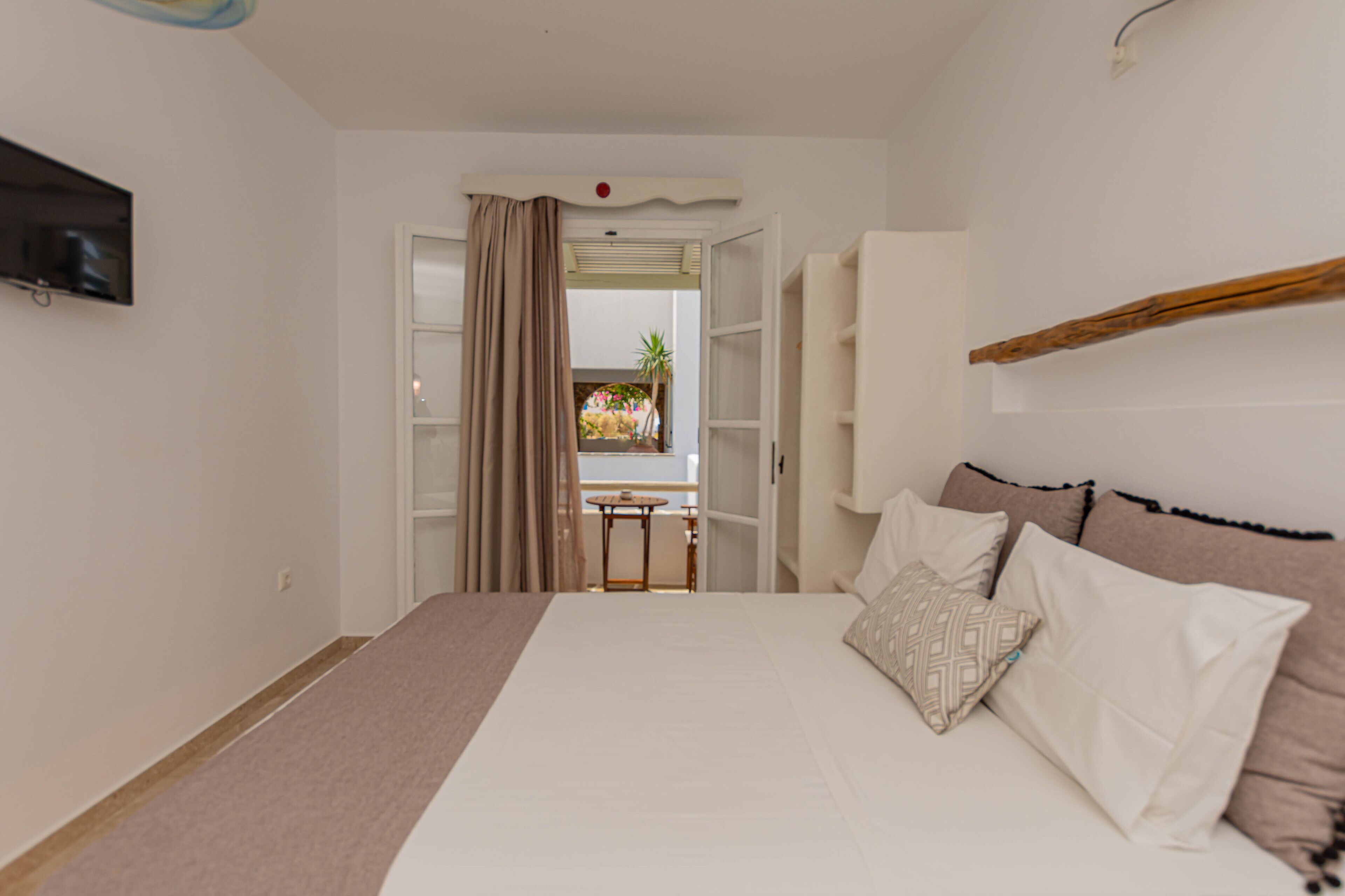 A bedroom with a bed, a TV mounted on the wall, and a view of a balcony with plants.