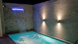 Ein rechteckiger Außenpool mit Wasserfall, umgeben von einer Ziegelmauer und zwei Wandleuchten.