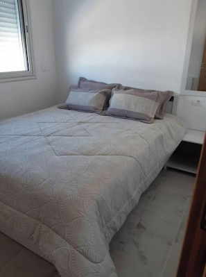 Un lit bien fait, recouvert d’une couvre-lit à motifs et agrémenté de deux oreillers, une table de chevet blanche et une fenêtre avec des stores.