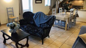Sala de estar com um sofá azul escuro, uma mesa de jantar e cadeiras. Há uma escada que leva a uma cozinha.
