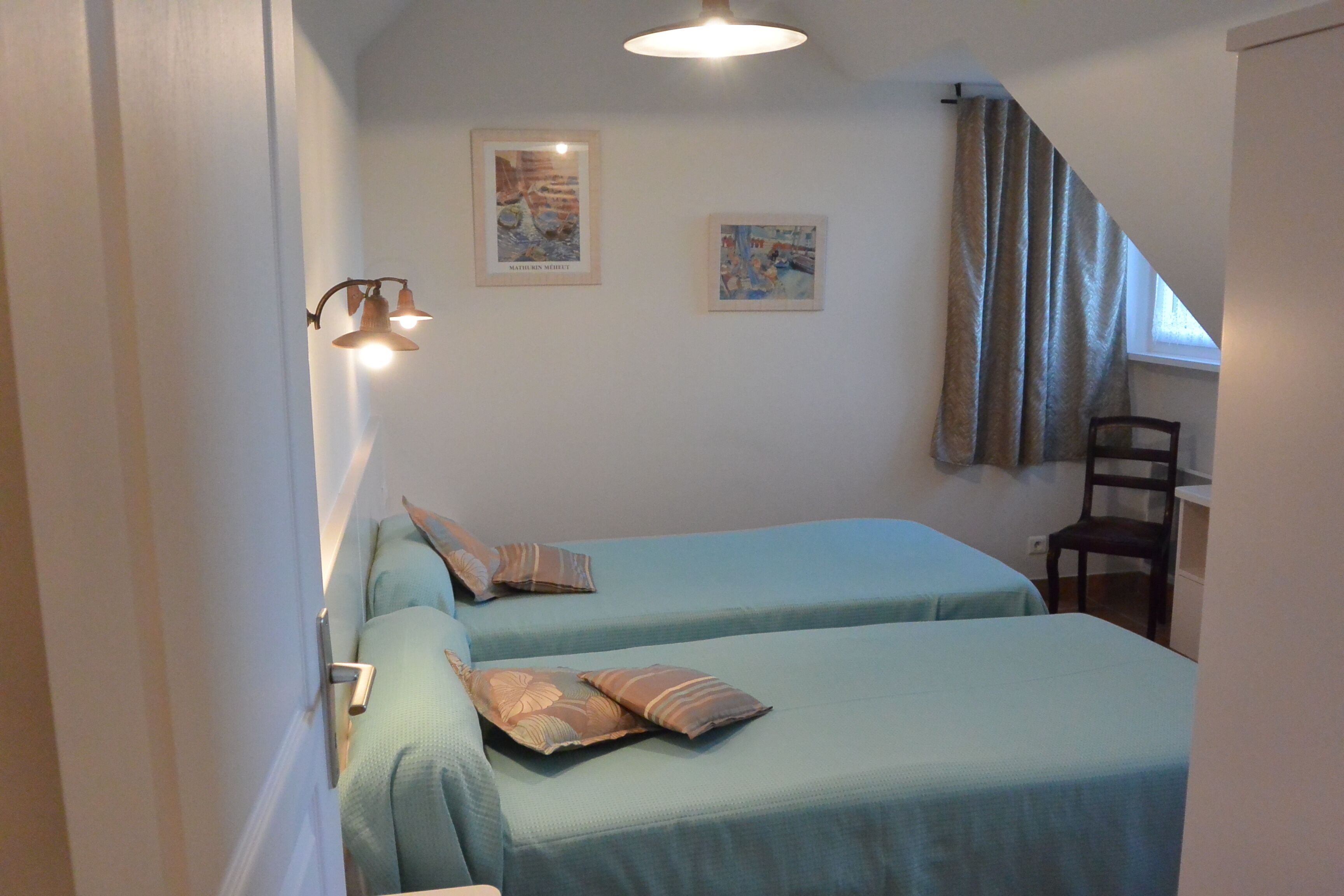 A room with two beds, a chair, a table, and two framed pictures on the wall.