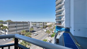 A high-rise building with multiple balconies, a street view with cars and palm trees, and a clear blue sky. - Spacious, Bay View - Close to Boardwalk, Location! (Myrtle Beach)