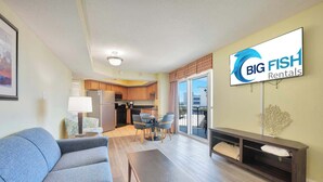 A living room with a sofa, a coffee table, and a TV mounted on the wall. There is a kitchen area with cabinets and a dining table. - Spacious, Bay View - Close to Boardwalk, Location! (Myrtle Beach)