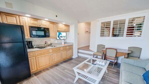 Una cocina compacta con armarios de madera, un refrigerador negro y un microondas. Hay un comedor con mesa y sillas, y un sofá en la sala.
