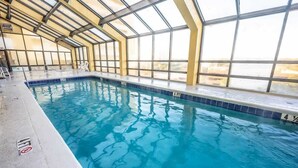 Une piscine intérieure avec des parois en verre transparent, un bord carrelé et un escalier menant à l’eau.