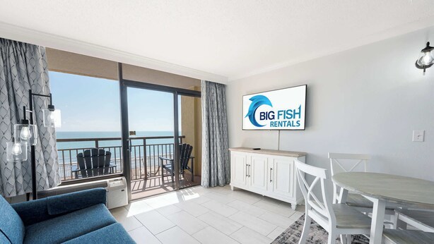 Un salon moderne avec balcon, un coin repas et un téléviseur diffusant des publicités pour « BIG FISH RENTALS ».