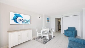 Un salon moderne avec un meuble blanc, une table à manger avec des chaises, un canapé bleu et un téléviseur affichant le logo « BIG FISH RENTALS ».