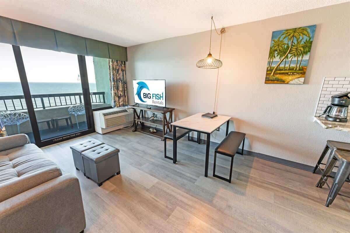 A modern living room with a large window overlooking the sea, a dining area with a table and chairs, a sofa, and a coffee station.