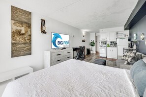 A modern bedroom with a large bed, a TV, and a dining area with a kitchen in the background.