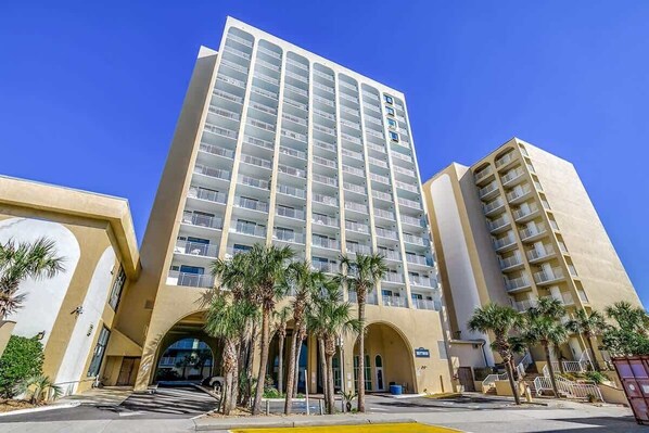 A high-rise hotel with multiple balconies, a central entrance, and palm trees in the foreground.