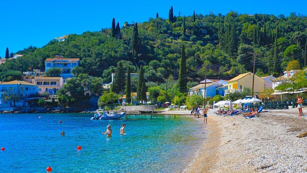 A beach with clear blue water, pebbly shore, and buildings in the background. - Agapi Studio in Kalami (Corfu)