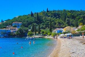 A beach with clear blue water, pebbly shore, and buildings in the background.