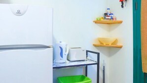 A compact kitchenette with a white refrigerator, a microwave, and a small shelf unit with a green basket and a white kettle. - Agapi Studio in Kalami (Corfu)