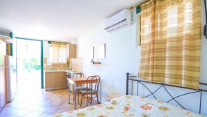 A bedroom with a bed, a desk, and a chair. There is a window with curtains, an air conditioning unit, and a view of the kitchen. - Agapi Studio in Kalami (Corfu)