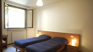 Ein Zimmer mit zwei Einzelbetten, einem Holzkopfstück, Nachttischlampen, einem Heizkörper und einem Fenster mit Blick auf Grünflächen.