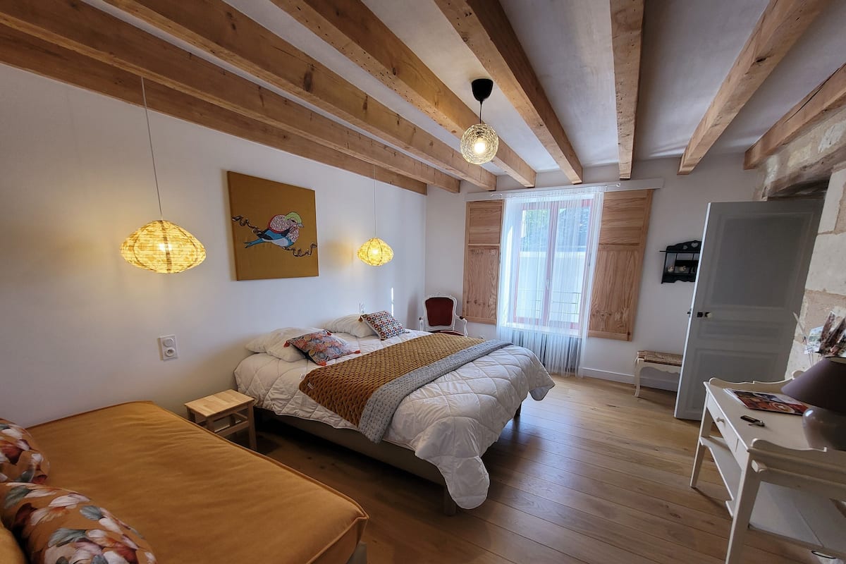 A bedroom with a wooden ceiling, a bed with a white duvet, a wooden bench, a white dresser, and a window with sheer curtains.