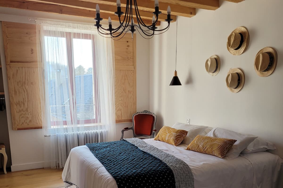 A bedroom with a bed, wooden floor, a chandelier, a window with curtains, and a wall with hats.