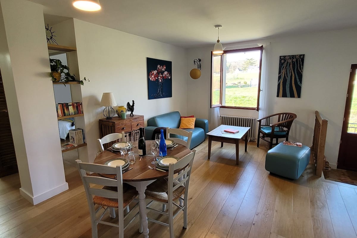 A spacious living room with a wooden floor, a dining area with a table set for four, a blue sofa, and a window providing natural light.