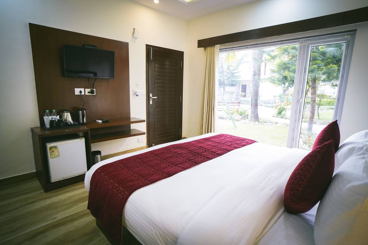 A hotel room with a bed, a TV mounted on the wall, a small refrigerator, and a window with a view of greenery.