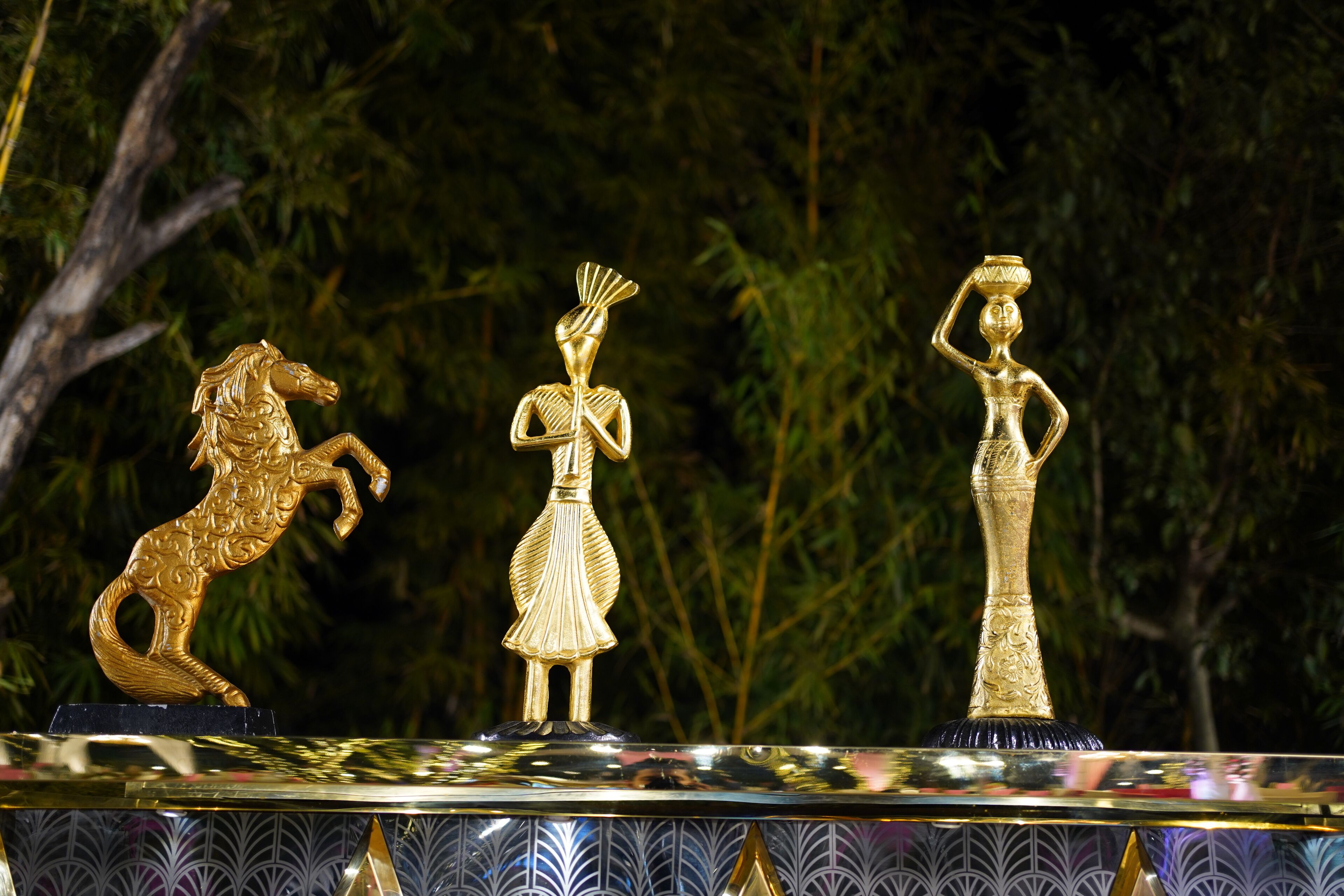 Tres estatuas doradas sobre una superficie reflectante con una base ornamentada, frente a un fondo de follaje denso.