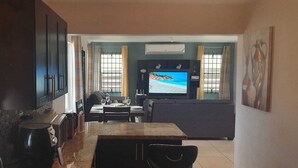 A dining area with a TV screen displaying a beach scene, a kitchen area with a stove, and a dining table with chairs. - Sea Front Villa  (Pool AC Gated 24hrs Security) (St. Catherine Parish Bridgeport P.O)