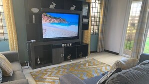 A living room with a flat-screen TV on a black entertainment unit, grey sofas, and a patterned rug. - Sea Front Villa  (Pool AC Gated 24hrs Security) (St. Catherine Parish Bridgeport P.O)