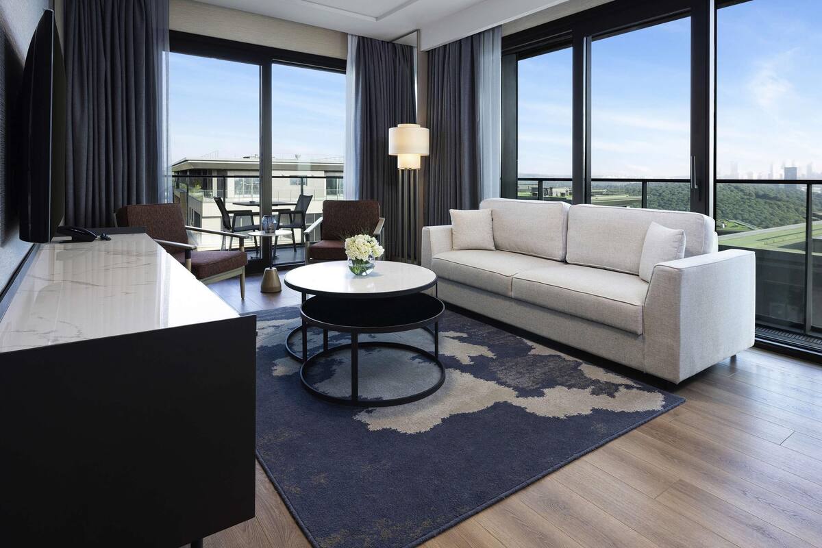 A modern hotel room with a large window, a sofa, a desk, and a dining area.