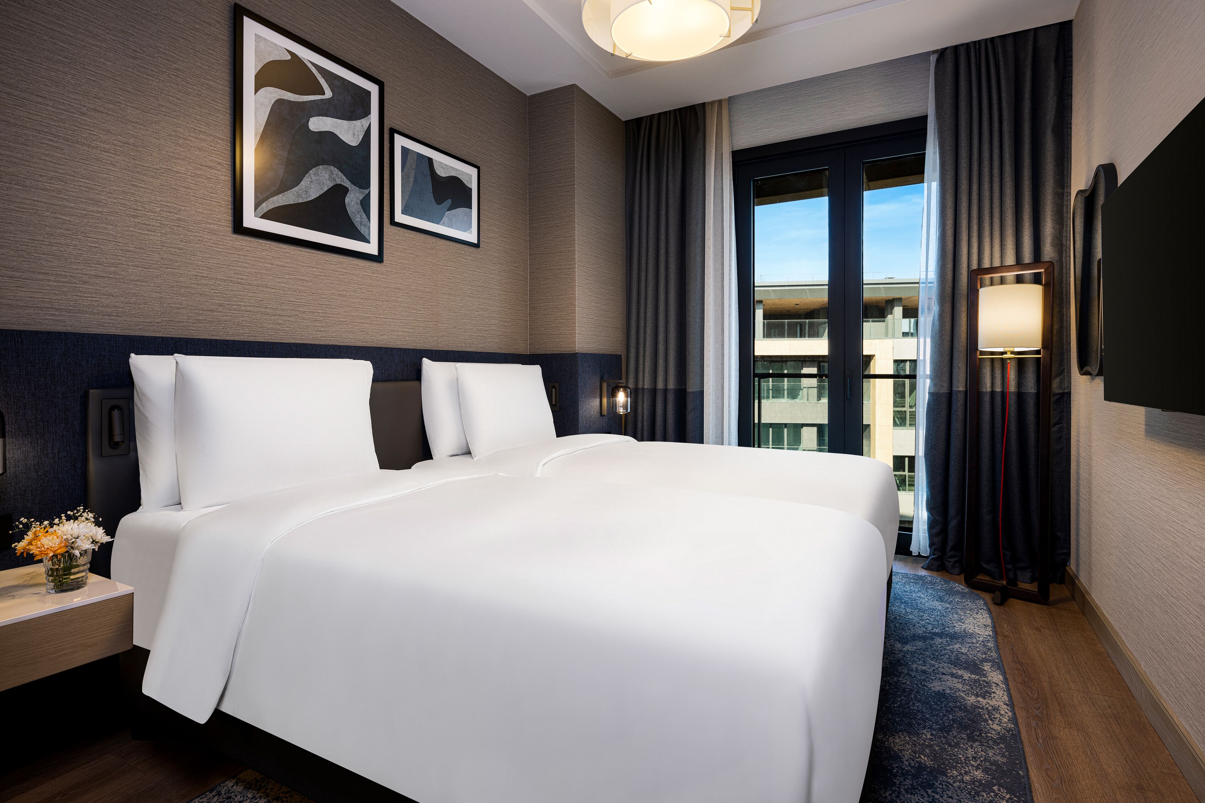 Habitación de hotel con una cama grande, una mesita de noche con un florero y un televisor montado en la pared, con vistas a la ciudad a través de la ventana.