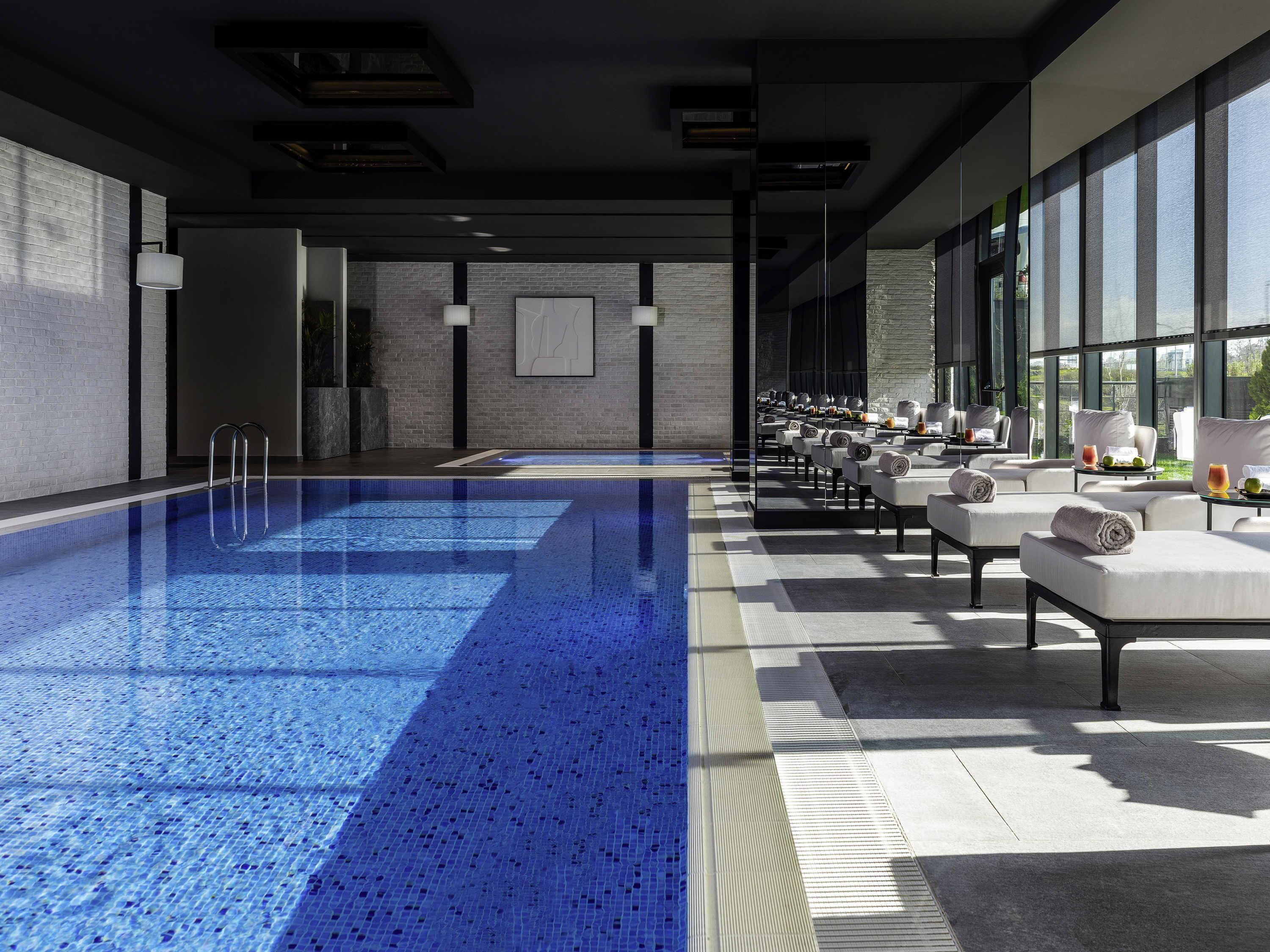 Una piscina interior moderna con superficie de agua azul cristalina, zona de estar contigua con asientos y grandes ventanales que ofrecen luz natural.