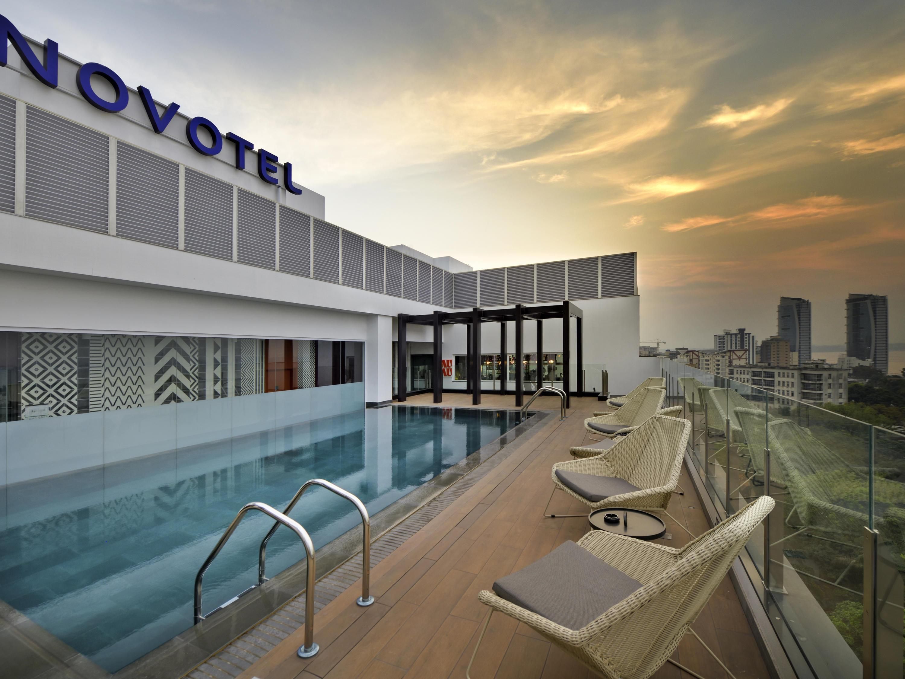 Une piscine sur le toit d’un hôtel Novotel, offrant une vue sur la ville, avec des chaises longues et un espace bar fermé par des parois de verre.