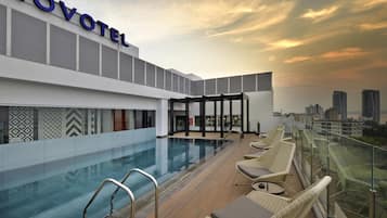 Une piscine sur le toit d’un hôtel Novotel, offrant une vue sur la ville, avec des chaises longues et un espace bar fermé par des parois de verre.